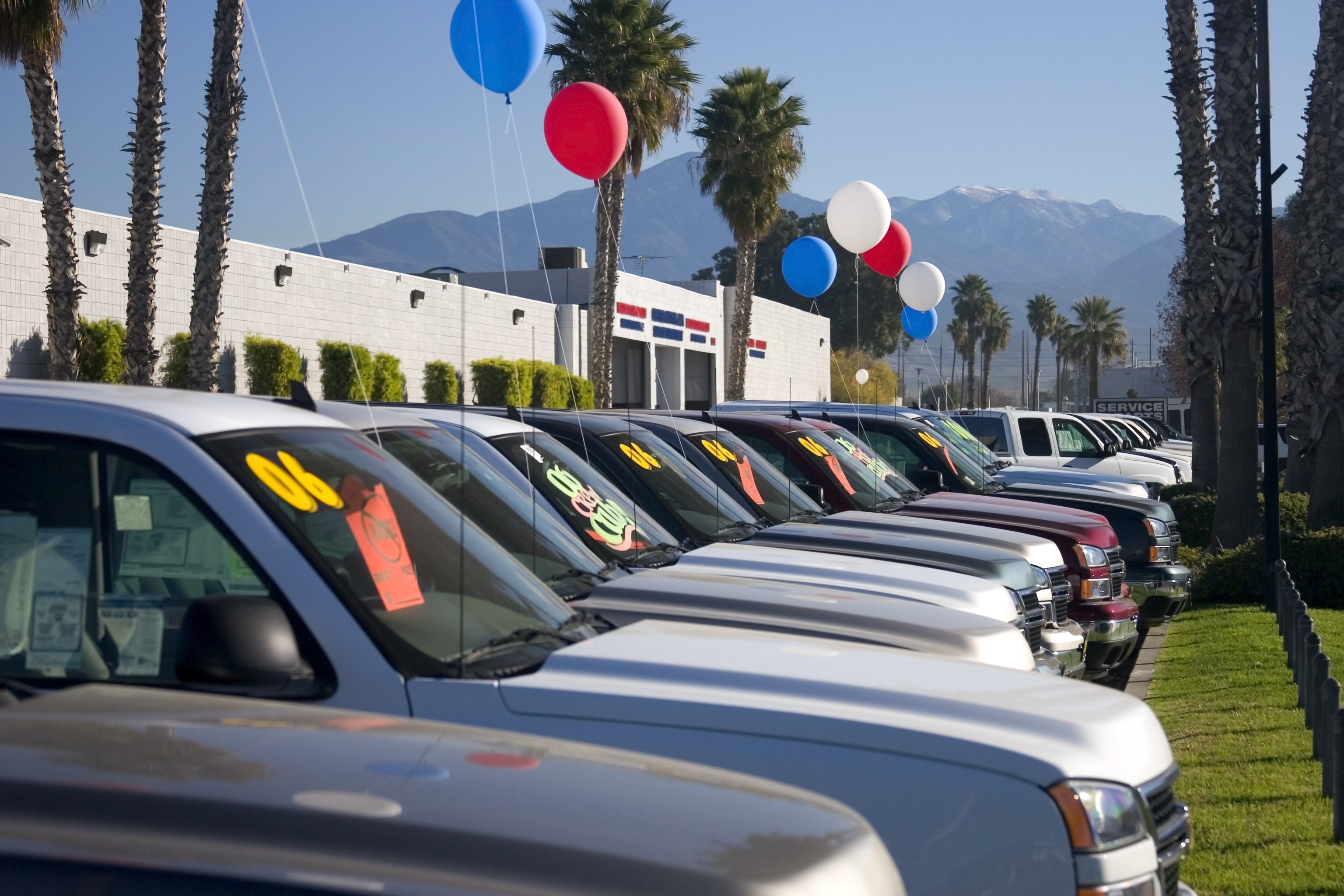 Row of used cars with sale signs in a car dealership lot with balloons and palm trees in the background.